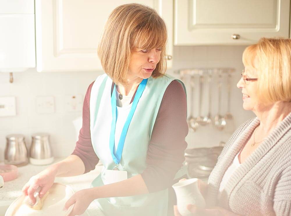 Caregiver washing up dishes and speaking to woman