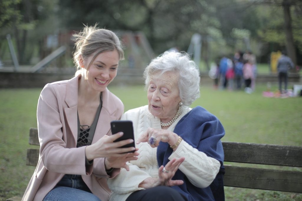 Younger lady showing an elderly lady something on her smart phone whilst they are sat outside on a bench in a park