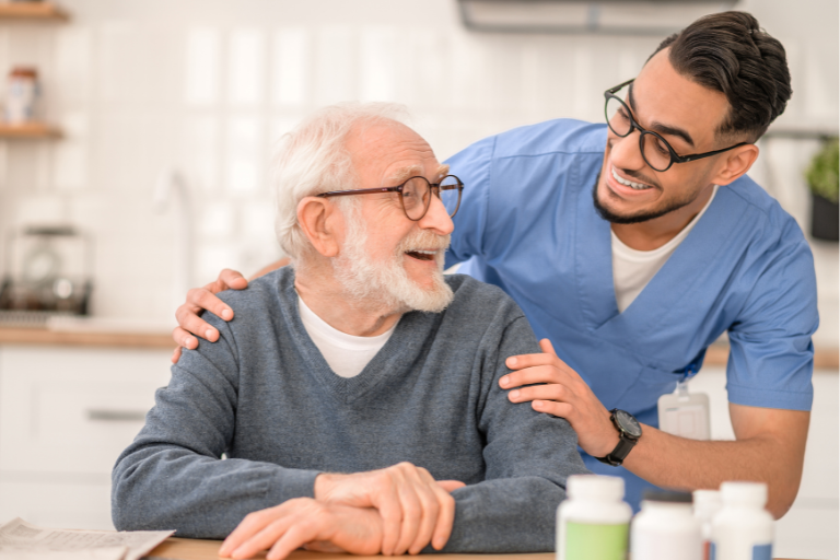 The Importance Of Consistency In Care: Why It Matters And How We Deliver It 1 Consistency In Care Shown As An Older Adult With White Hair And Glasses Seated At A Table, While A Healthcare Professional In Blue Medical Scrubs Stands Nearby With A Hand Resting On The Seated Person’s Shoulder; Several Medication Bottles Are Arranged On The Table In A Bright Kitchen Setting.