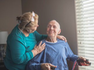 Older adult seated near a window holding a walking aid, with another older adult standing beside them and placing a hand on the seated person’s shoulder inside a softly lit room.